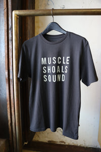 A black t-shirt hanging on a hanger from a brass clothing rack. The shirt features the text 'MUSCLE SHOALS SOUND' printed in a bold, white sans-serif font across the chest.