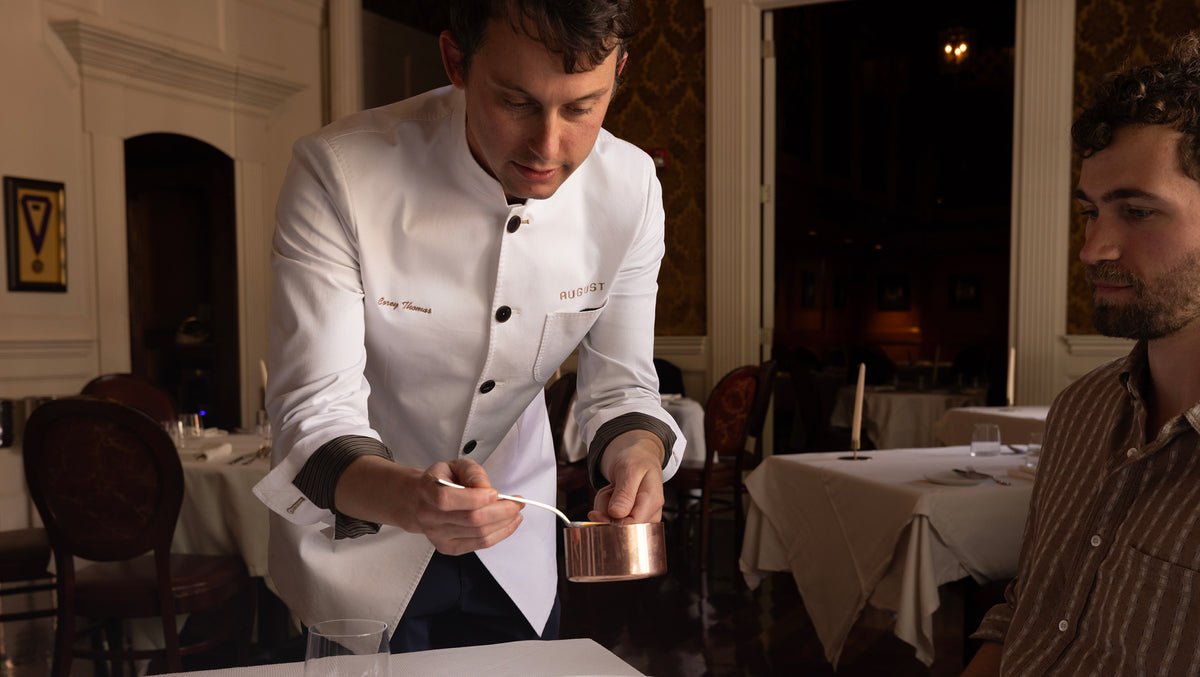A chef in a white uniform with black trim holds a small copper measuring cup and spoon in a restaurant setting, with another man partially visible on the right.