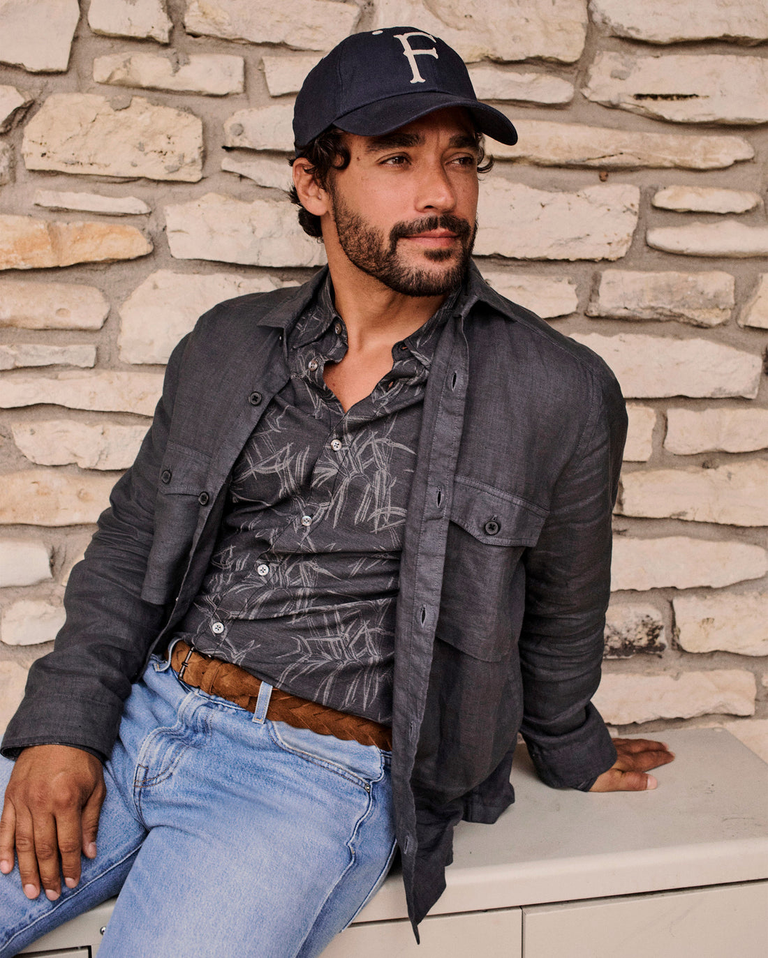 Man wearing a cap and dark jacket sitting against a stone wall.