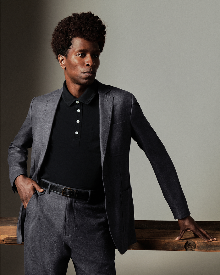 Man wearing a gray blazer over a black shirt with white buttons, standing against a neutral background.