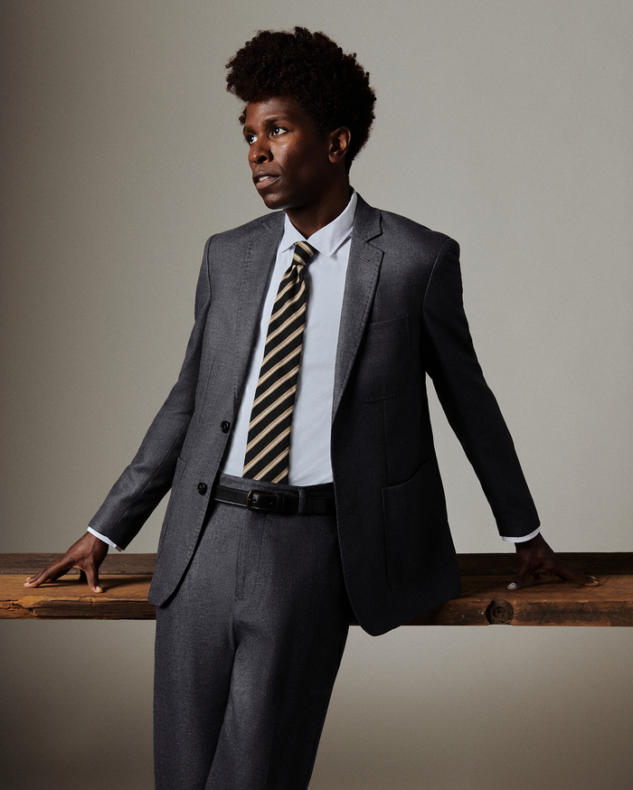 Man in a gray suit with a striped tie leaning against a wooden surface.