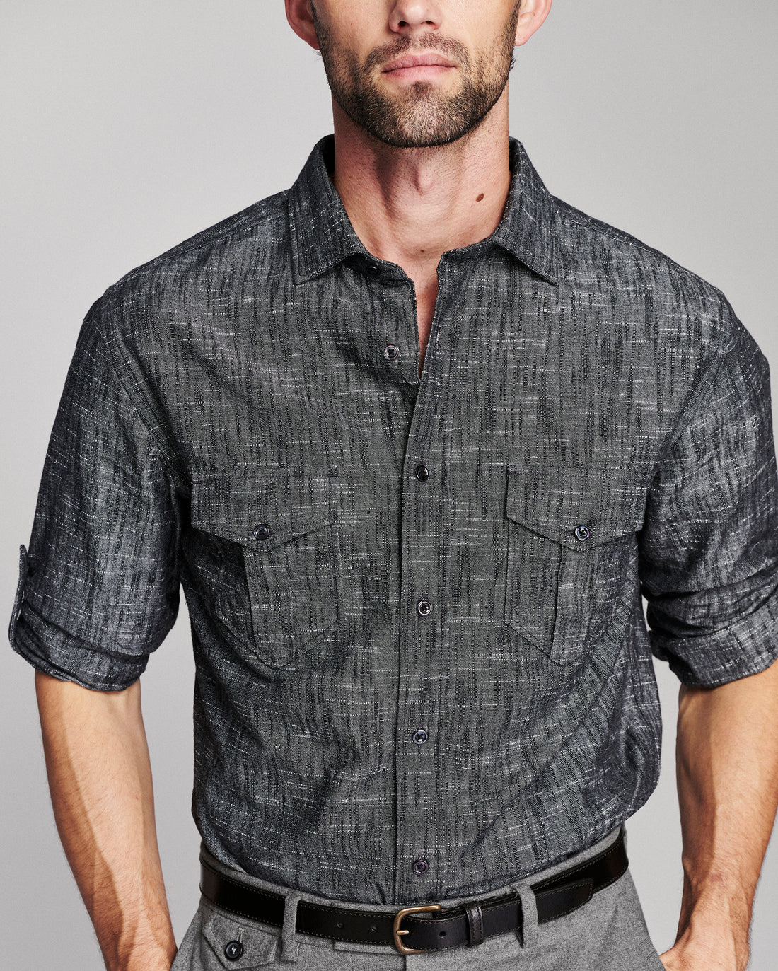 A man wearing a dark gray textured short-sleeve button-up shirt with two chest pockets, paired with gray trousers and a black belt, against a light gray studio background.