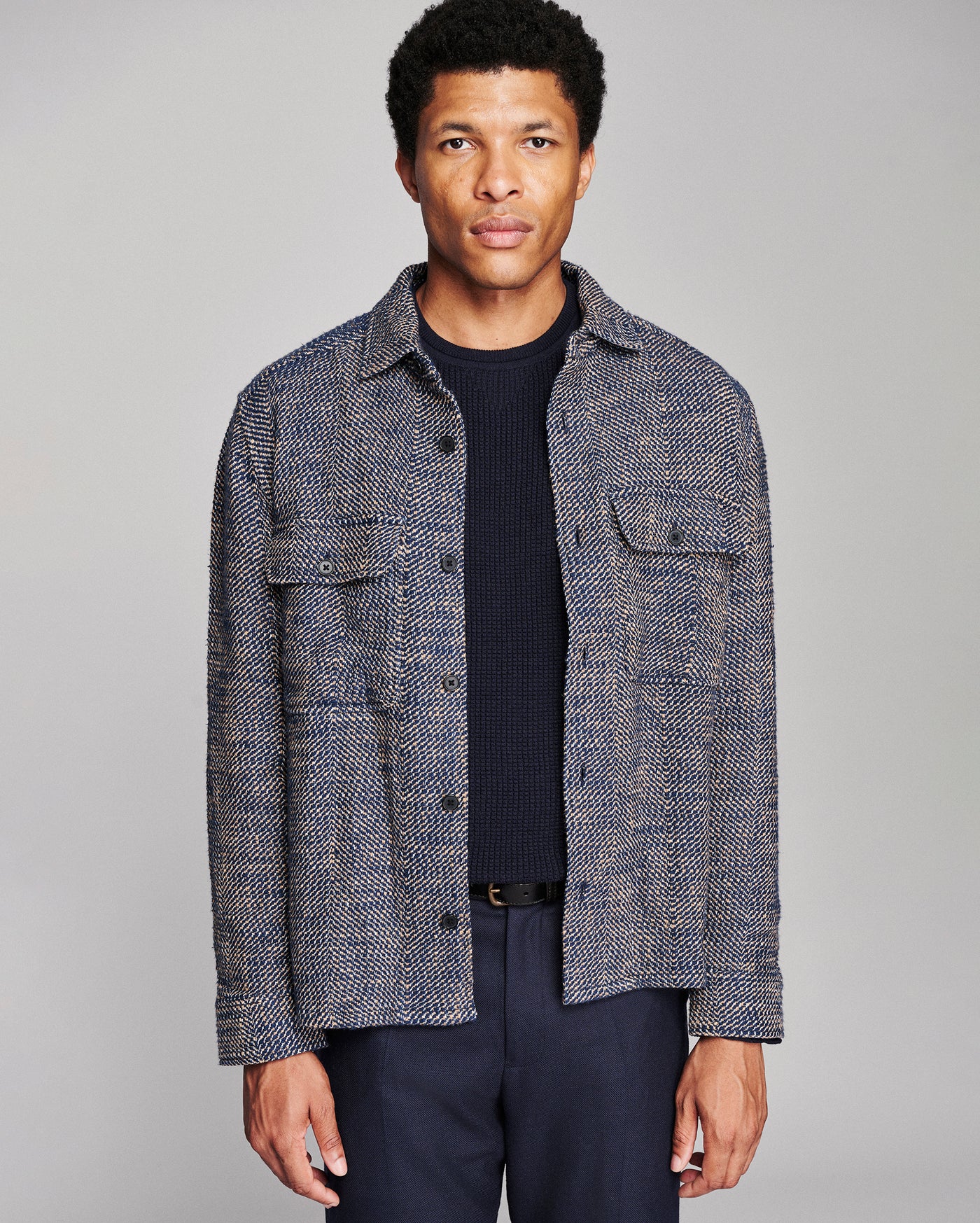 A man wearing a textured gray and navy tweed button-up shirt jacket over a dark navy ribbed sweater and navy trousers, photographed against a solid light gray studio background.