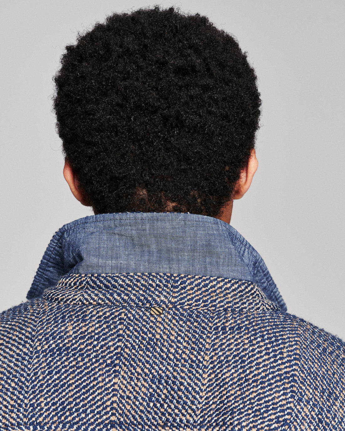 Back view of a person with short, curly black hair wearing a textured blue and beige herringbone jacket against a solid gray background.
