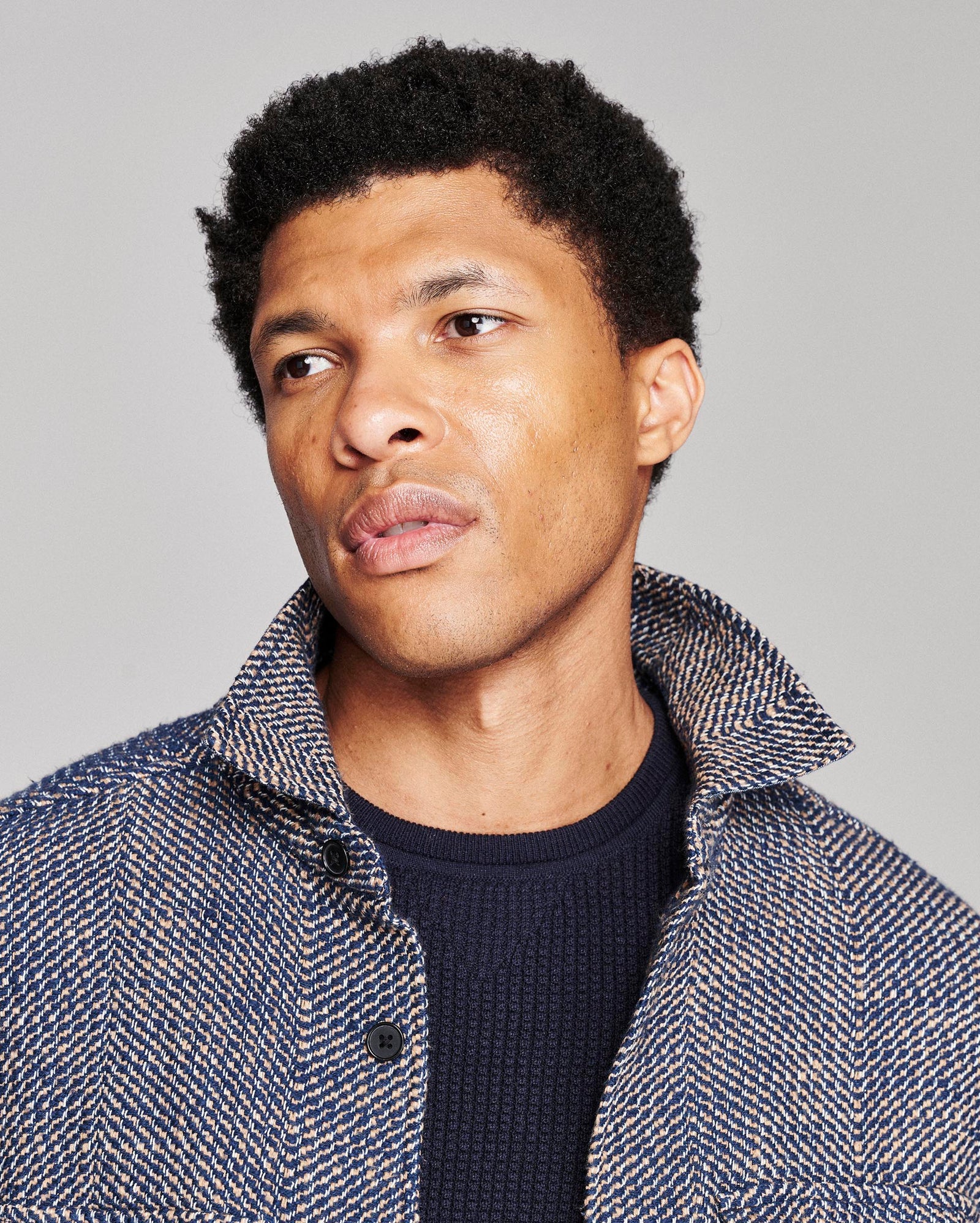 A close-up portrait of a man with short black curly hair, wearing a textured blue and white herringbone jacket over a dark navy knit shirt, set against a solid light gray background.