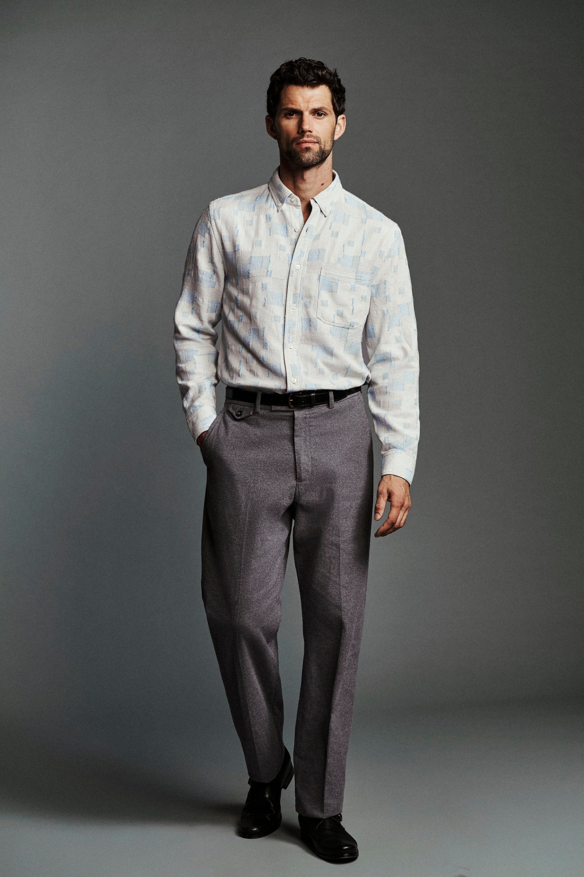 A man modeling a light-colored, subtly patterned long-sleeve button-down shirt paired with gray trousers, a black belt, and black dress shoes against a dark gray studio background.