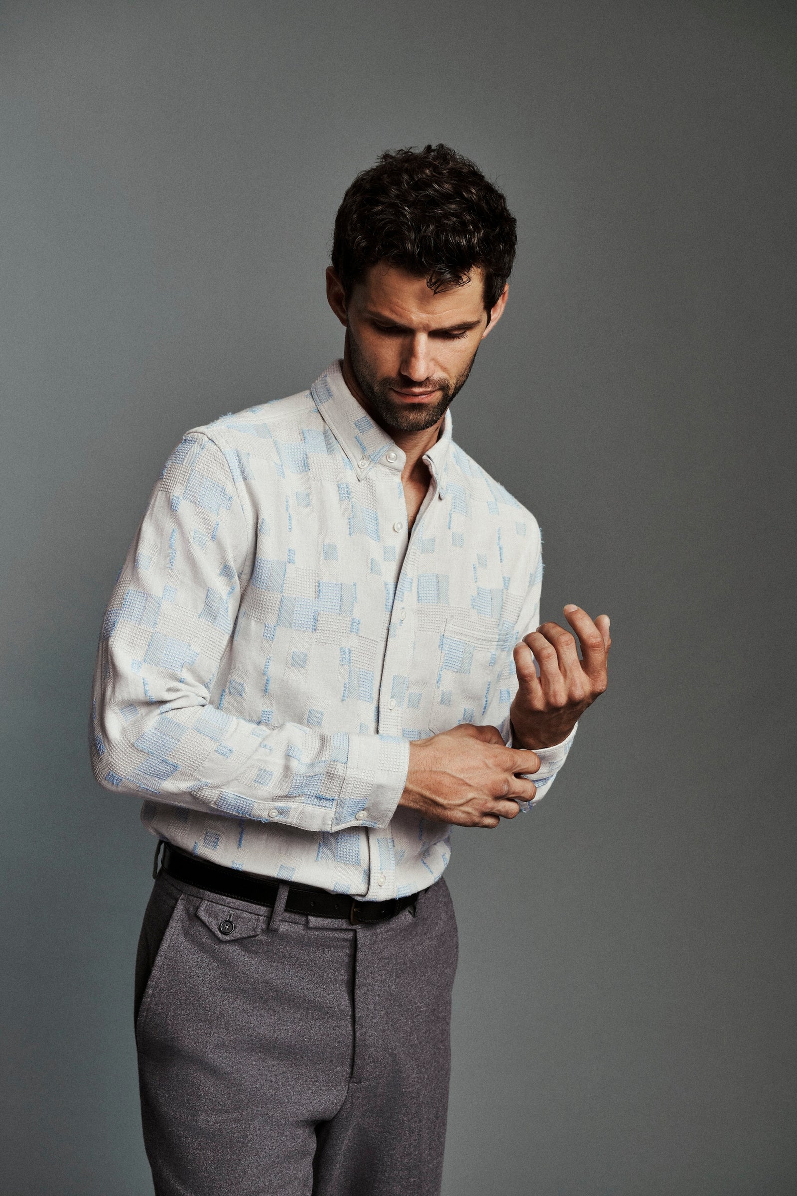 A man wearing a light blue and white patterned button-down shirt and gray trousers, adjusting his shirt cuff against a solid gray studio background.