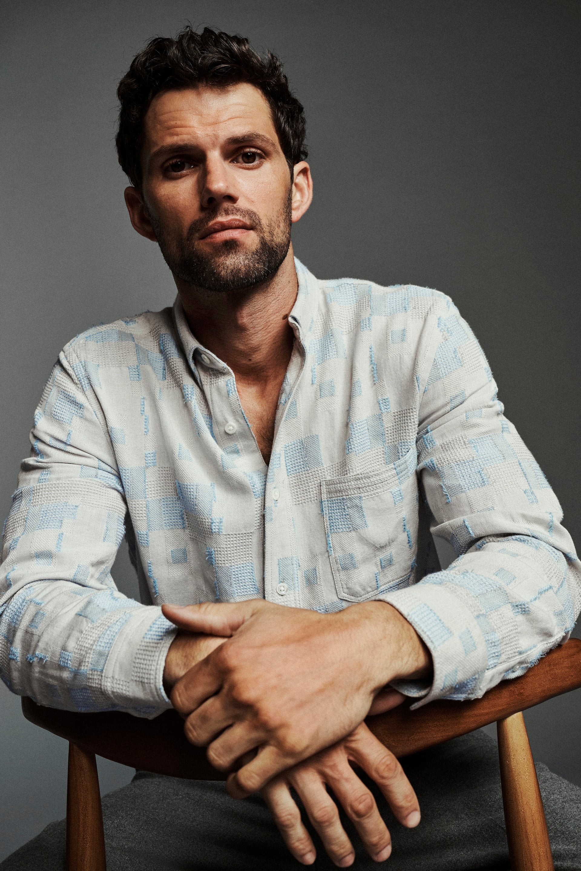 A man wearing a light blue and white patterned button-down shirt, sitting on a wooden chair against a dark gray studio background.