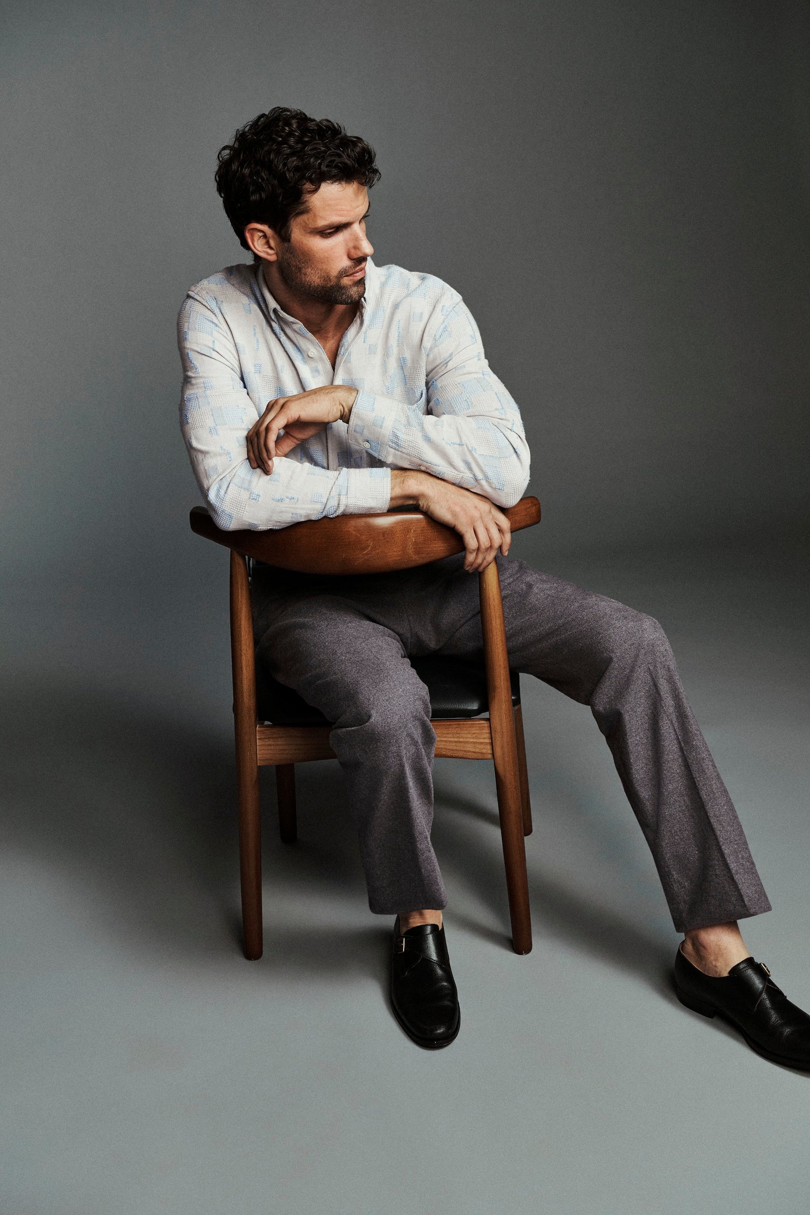 A man sits on a wooden chair in a studio setting, wearing a light-colored long-sleeve button-up shirt, gray trousers, and black dress shoes against a solid gray background.