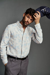 A man wearing a light blue and white patterned button-down shirt, gray trousers, and a navy blue cap with a white logo, posed against a solid gray studio background.