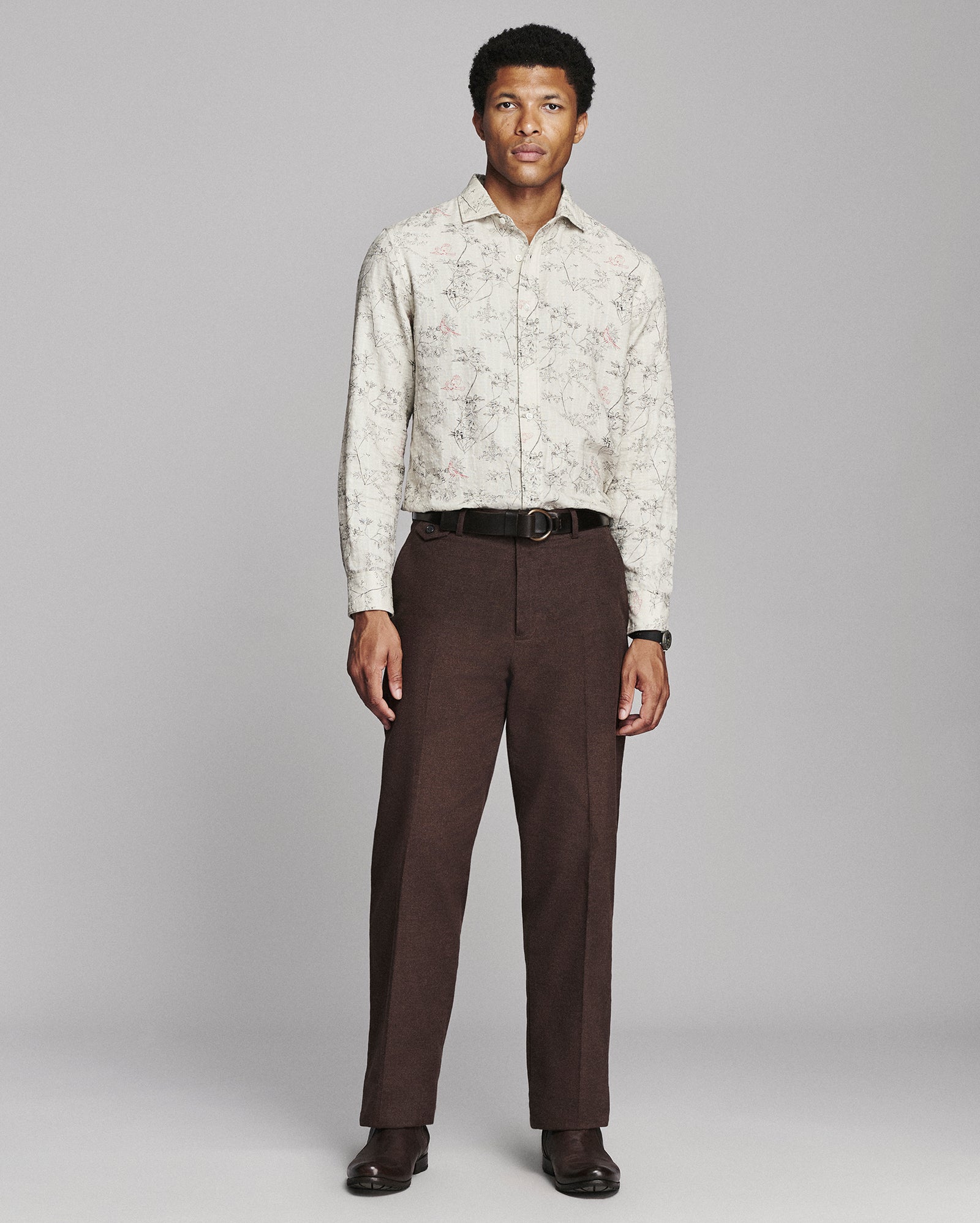A man modeling a long-sleeve button-up shirt with a light beige and brown abstract pattern, paired with dark brown trousers, a black belt, and dark brown dress shoes, against a solid gray studio background.
