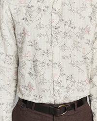 A close-up of a long-sleeve button-up shirt with a delicate botanical and bird print on a light beige fabric, styled with a dark brown belt and dark brown trousers.