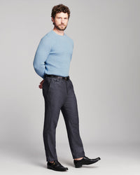A man modeling a light blue crewneck sweater and dark charcoal gray trousers with black leather dress shoes, standing against a light gray studio background.