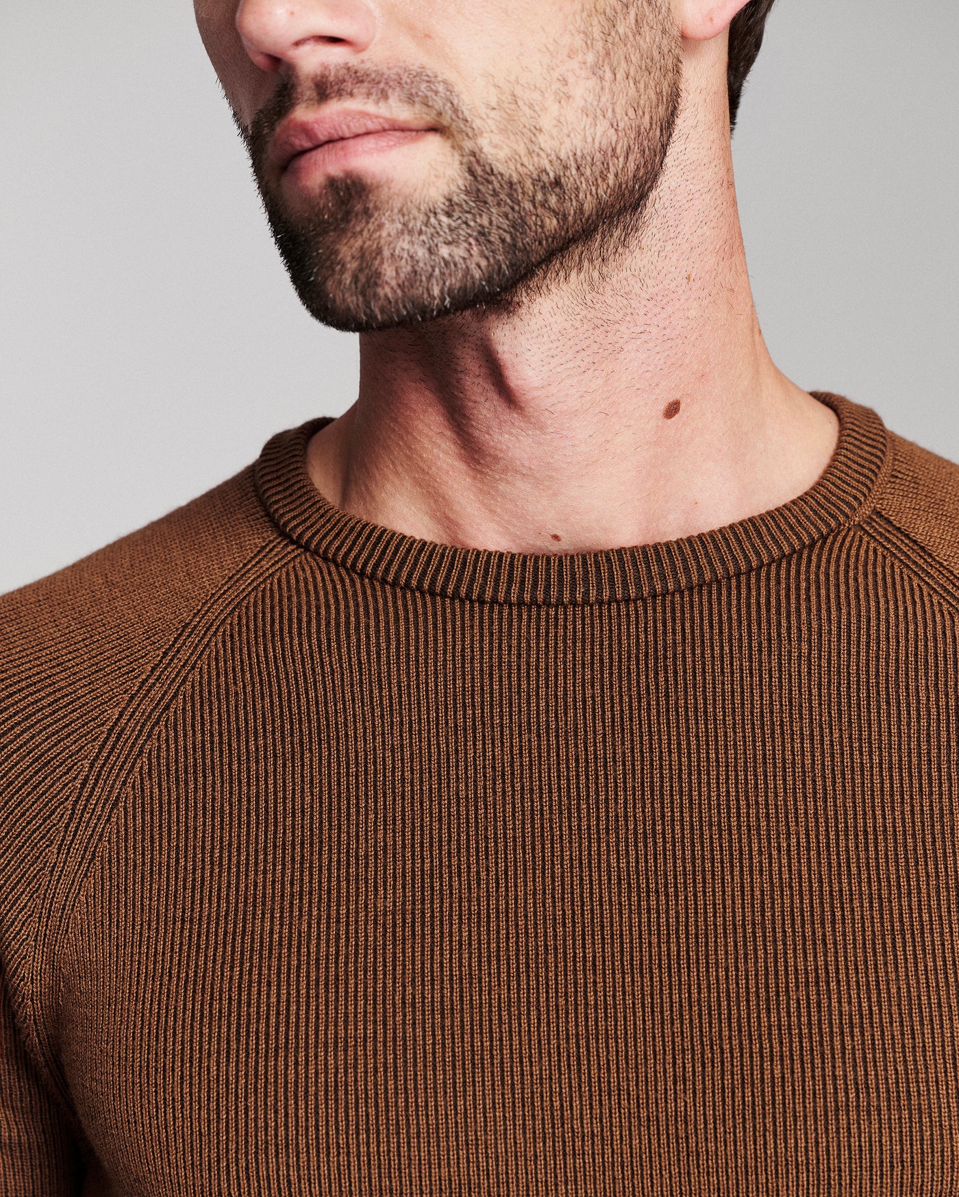 Close-up of a man wearing a brown ribbed crewneck sweater, showing the texture of the knit fabric and the collar.
