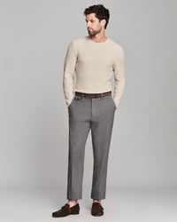 A man modeling a light beige knit crewneck sweater, gray trousers, a brown belt, and dark brown loafers against a solid light gray studio background.