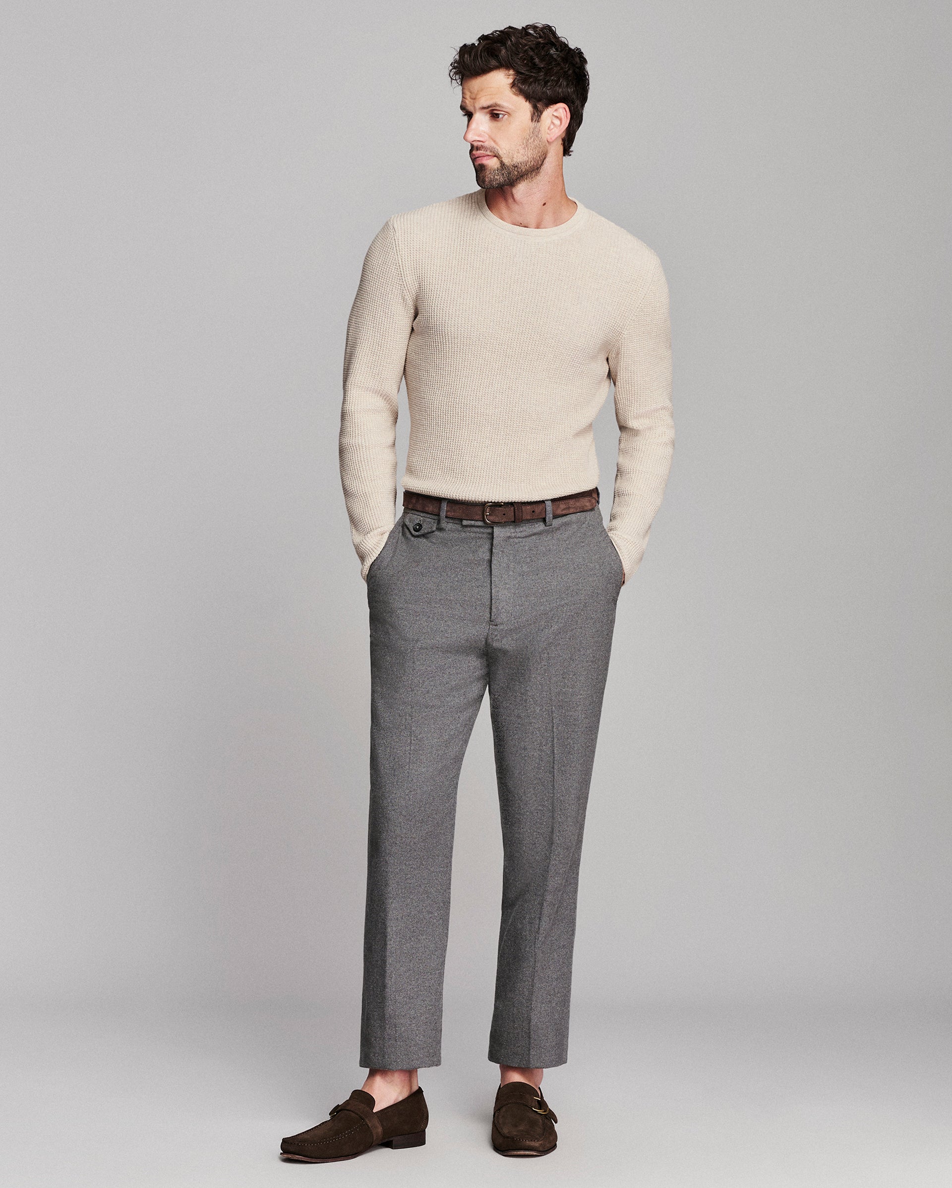 A man modeling a light beige knit crewneck sweater, gray trousers, a brown belt, and dark brown loafers against a solid light gray studio background.