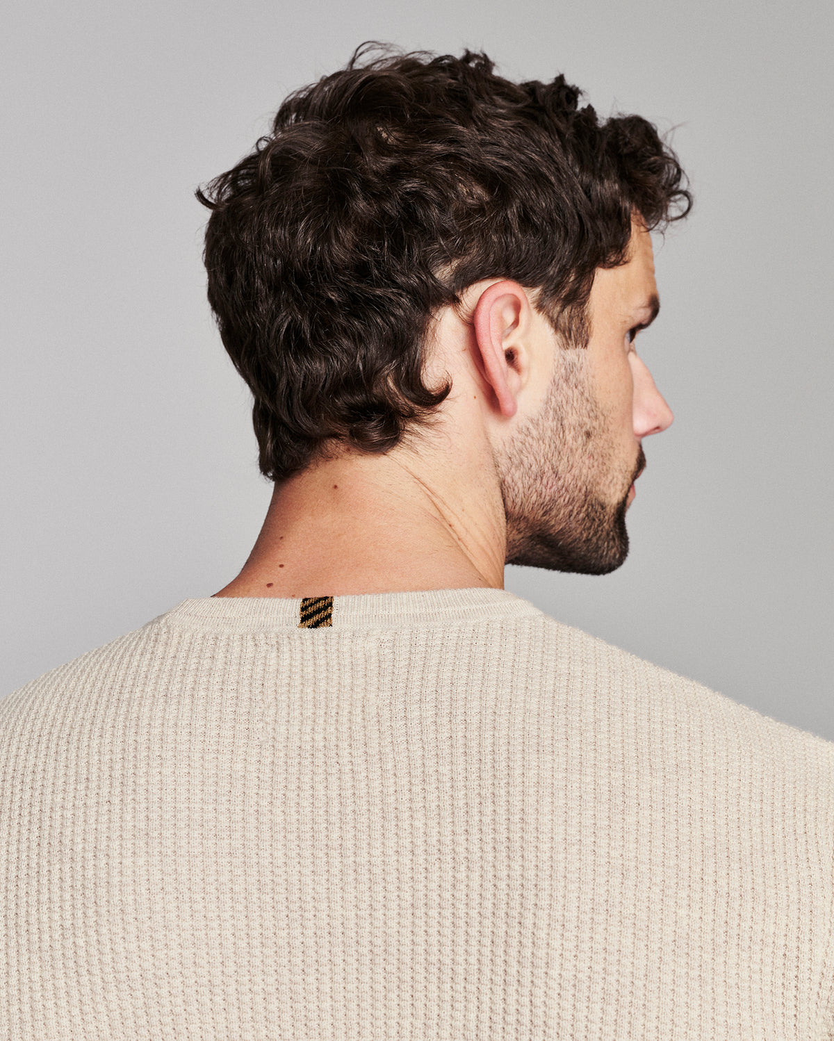 A man shown from the back and side profile wearing a cream-colored textured knit sweater against a solid gray background.