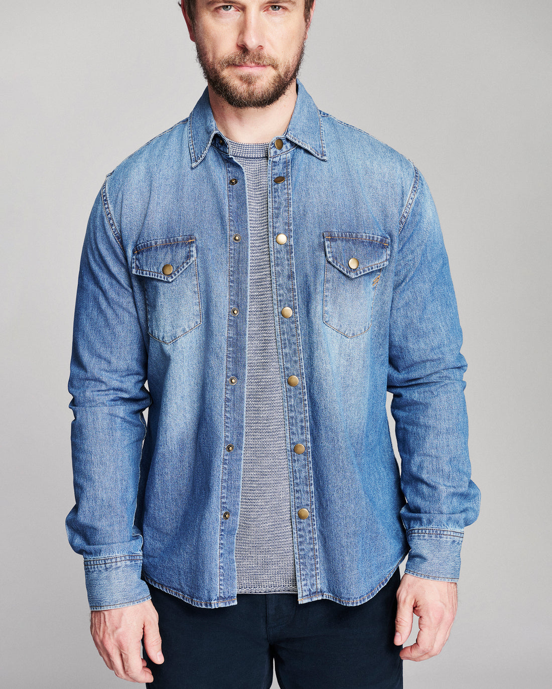 A man wearing a light blue denim button-up shirt over a gray striped t-shirt and dark pants, photographed against a solid light gray studio background.