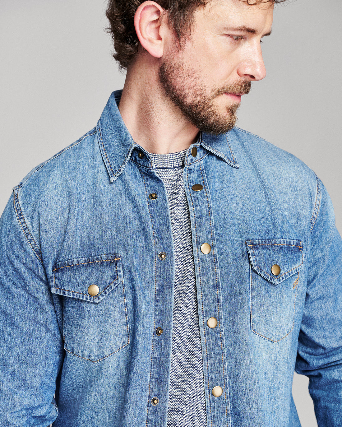 A man wearing a light blue denim button-up shirt over a gray knit crewneck sweater, shown in a close-up profile view against a solid gray background.
