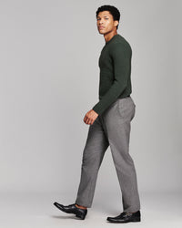 A man in a dark green long-sleeve knit sweater and gray trousers walking in profile against a light gray studio background.