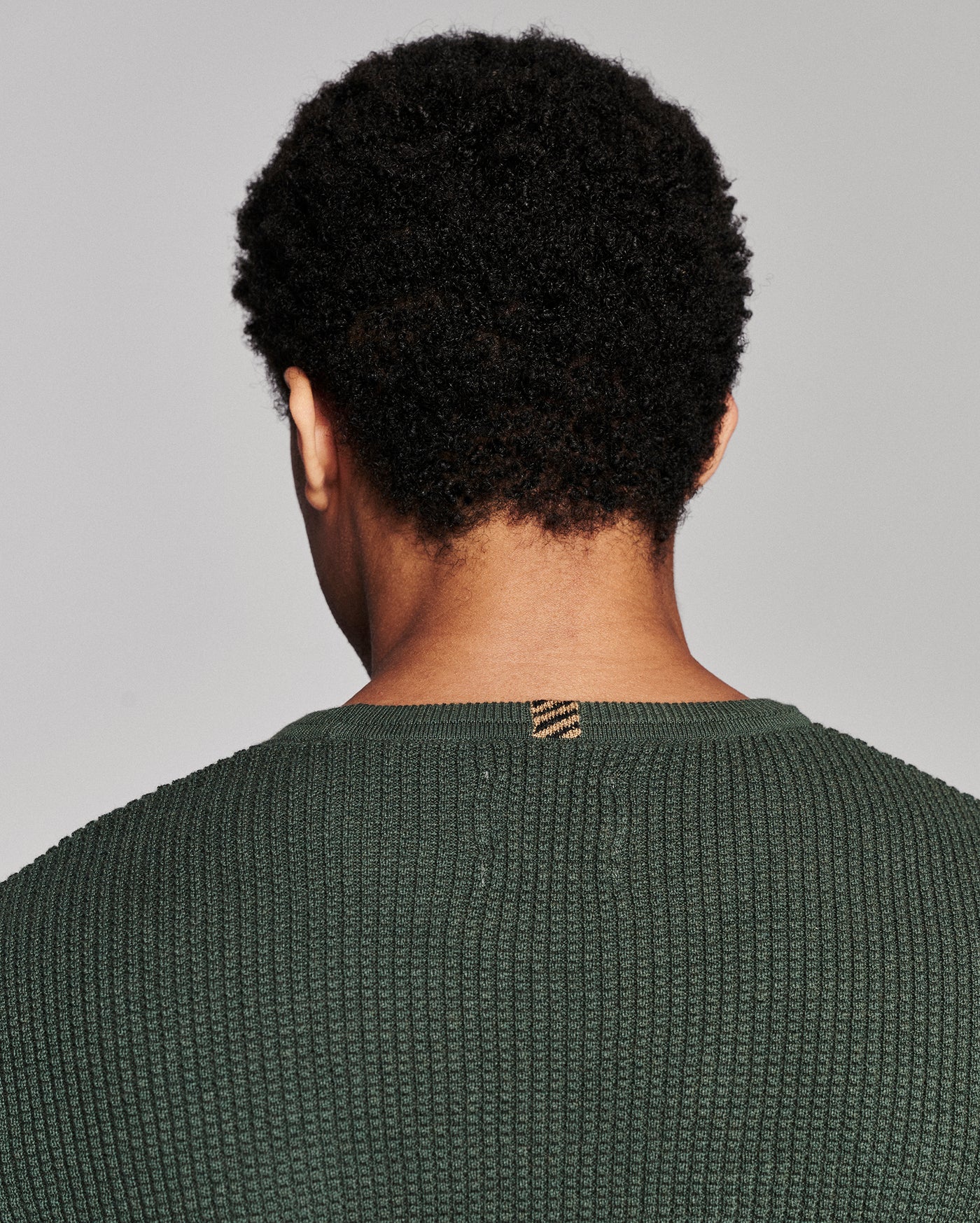 Back view of a person with short curly black hair wearing a textured dark green knit sweater against a light gray studio background.