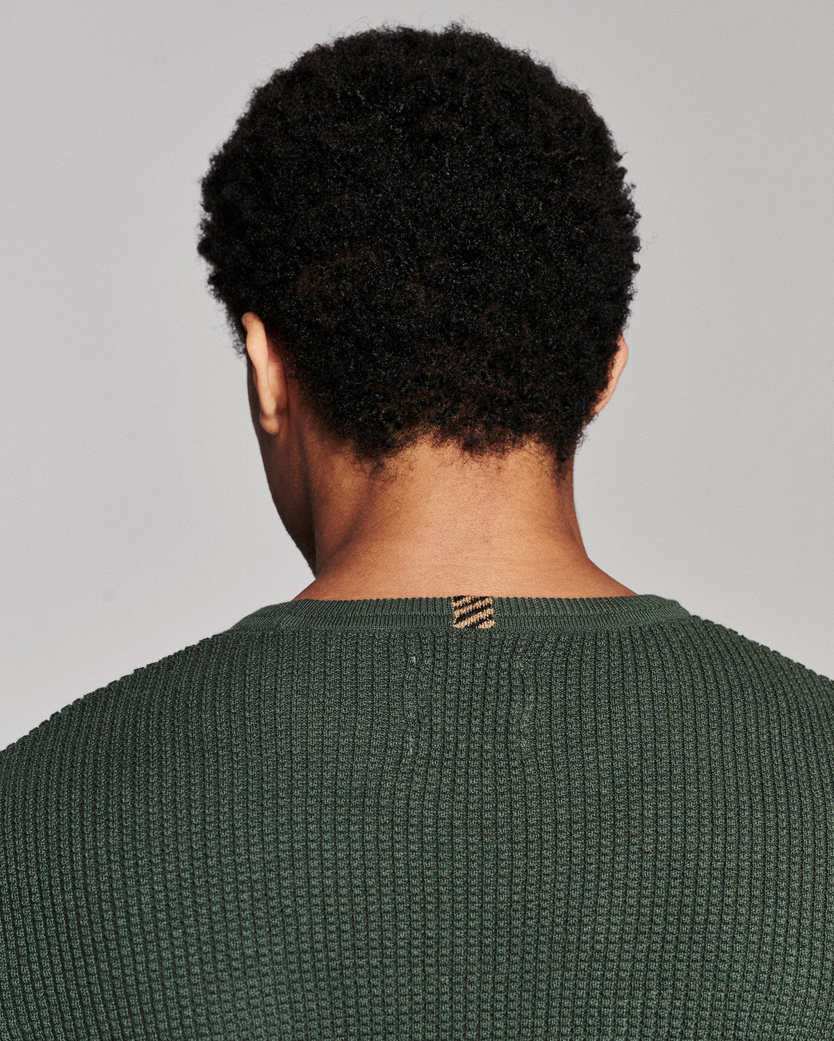 Back view of a person with short curly black hair wearing a textured dark green knit sweater against a light gray studio background.