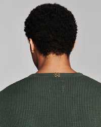 Back view of a person with short curly black hair wearing a textured dark green knit sweater against a light gray studio background.