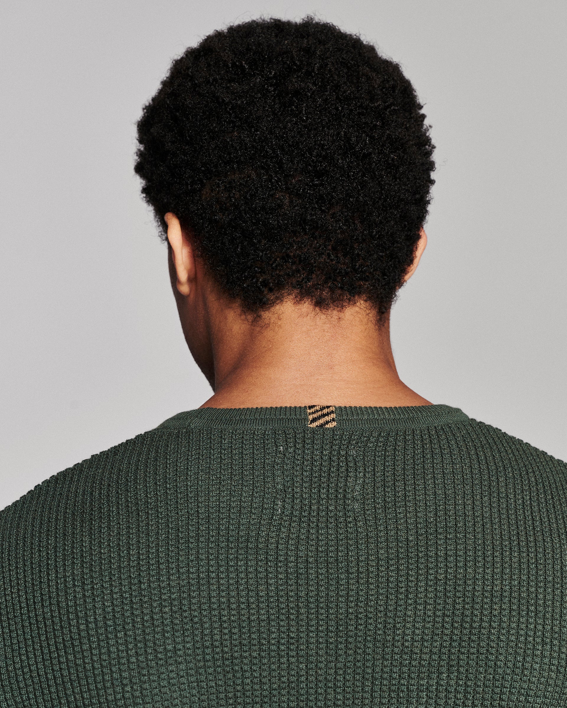 Back view of a person with short curly black hair wearing a textured dark green knit sweater against a light gray studio background.
