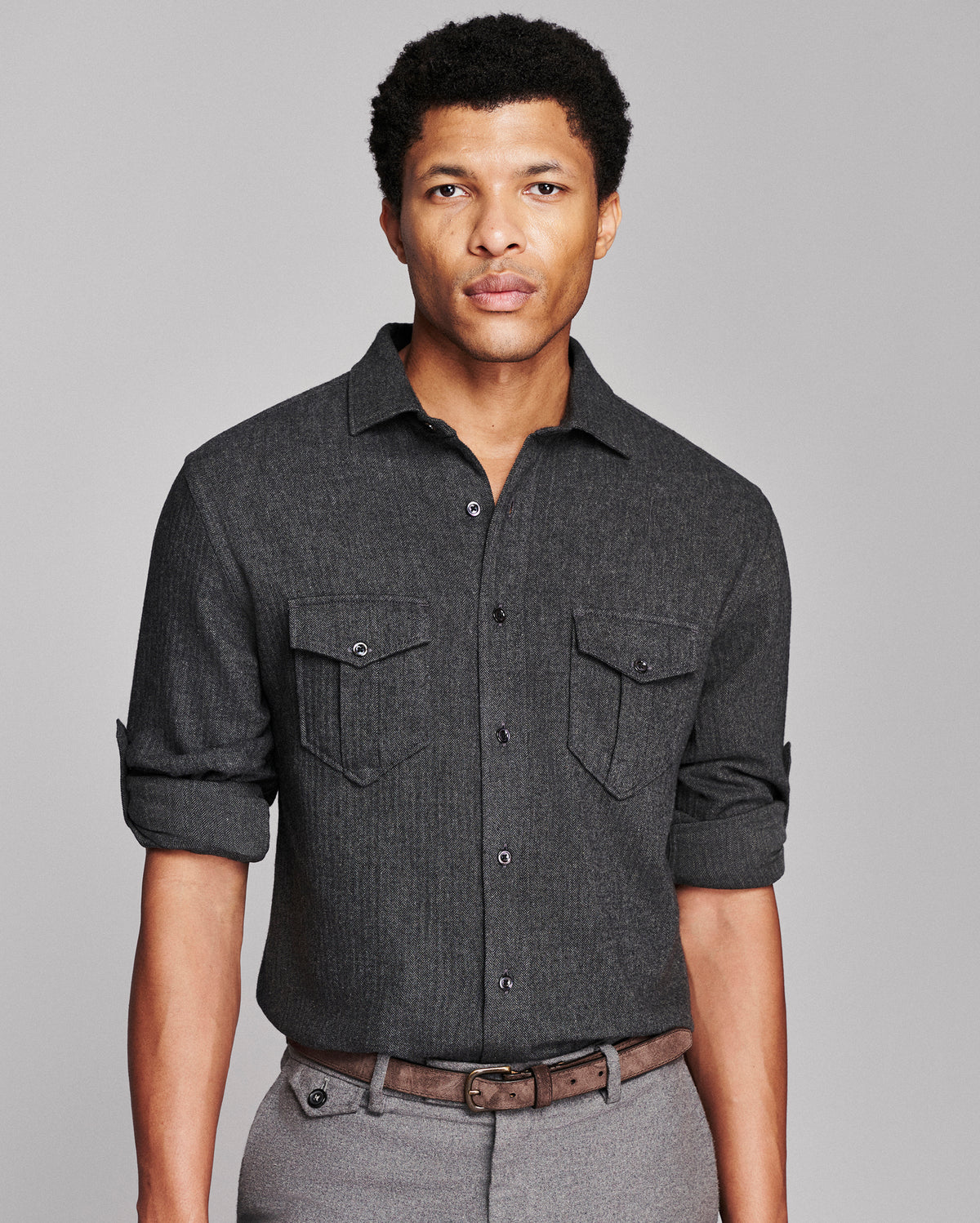 A man wearing a dark charcoal gray textured button-down shirt with two chest pockets, paired with gray trousers and a brown belt, against a solid gray studio background.