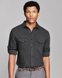 A man wearing a dark charcoal gray textured button-down shirt with two chest pockets, paired with gray trousers and a brown belt, against a solid gray studio background.