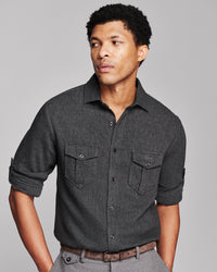 A man wearing a dark charcoal gray textured long-sleeve button-down shirt with two chest pockets, paired with gray trousers and a brown belt, photographed against a solid gray studio background.