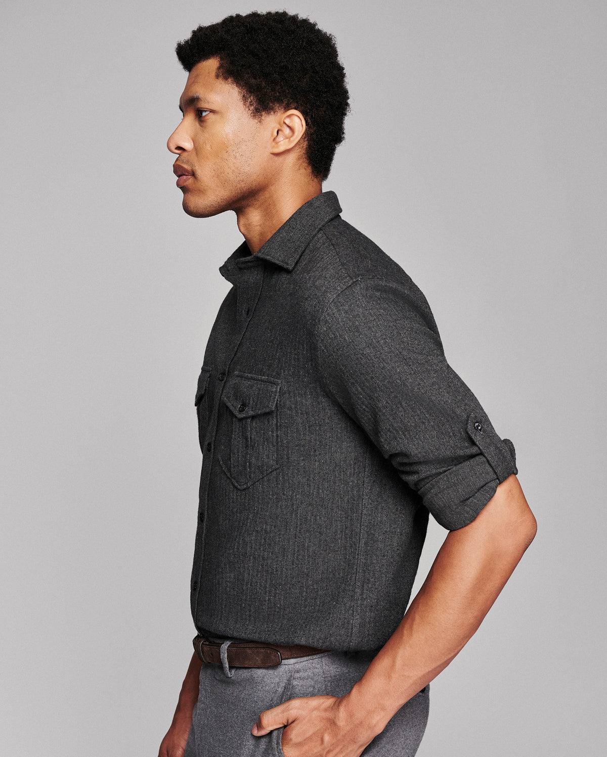 A man in profile wearing a dark charcoal gray textured button-down shirt with rolled-up sleeves and gray trousers, photographed against a solid light gray studio background.