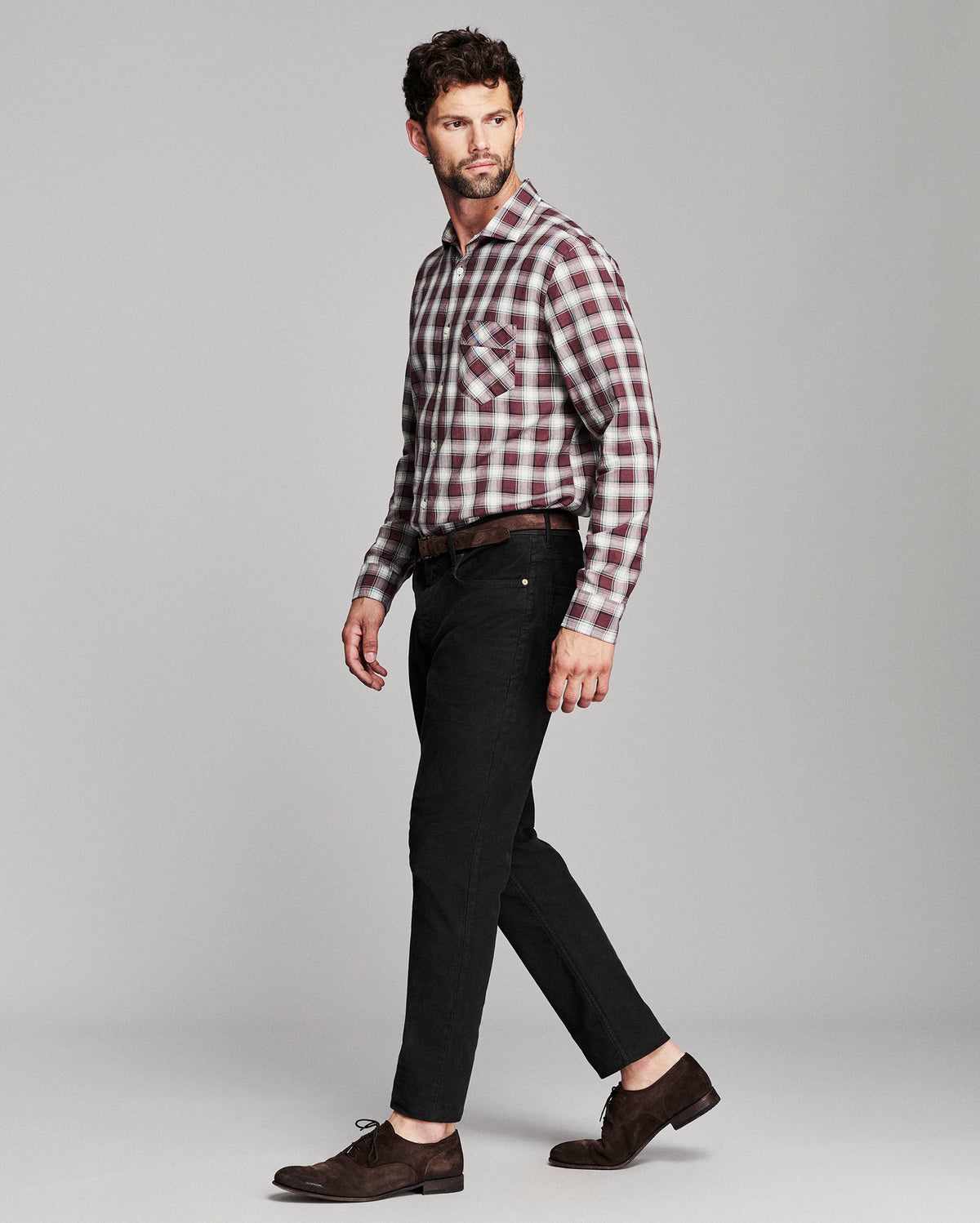 A man modeling a long-sleeve red and white plaid button-down shirt, paired with black jeans, a dark brown belt, and dark brown suede oxford shoes, against a solid light gray background.