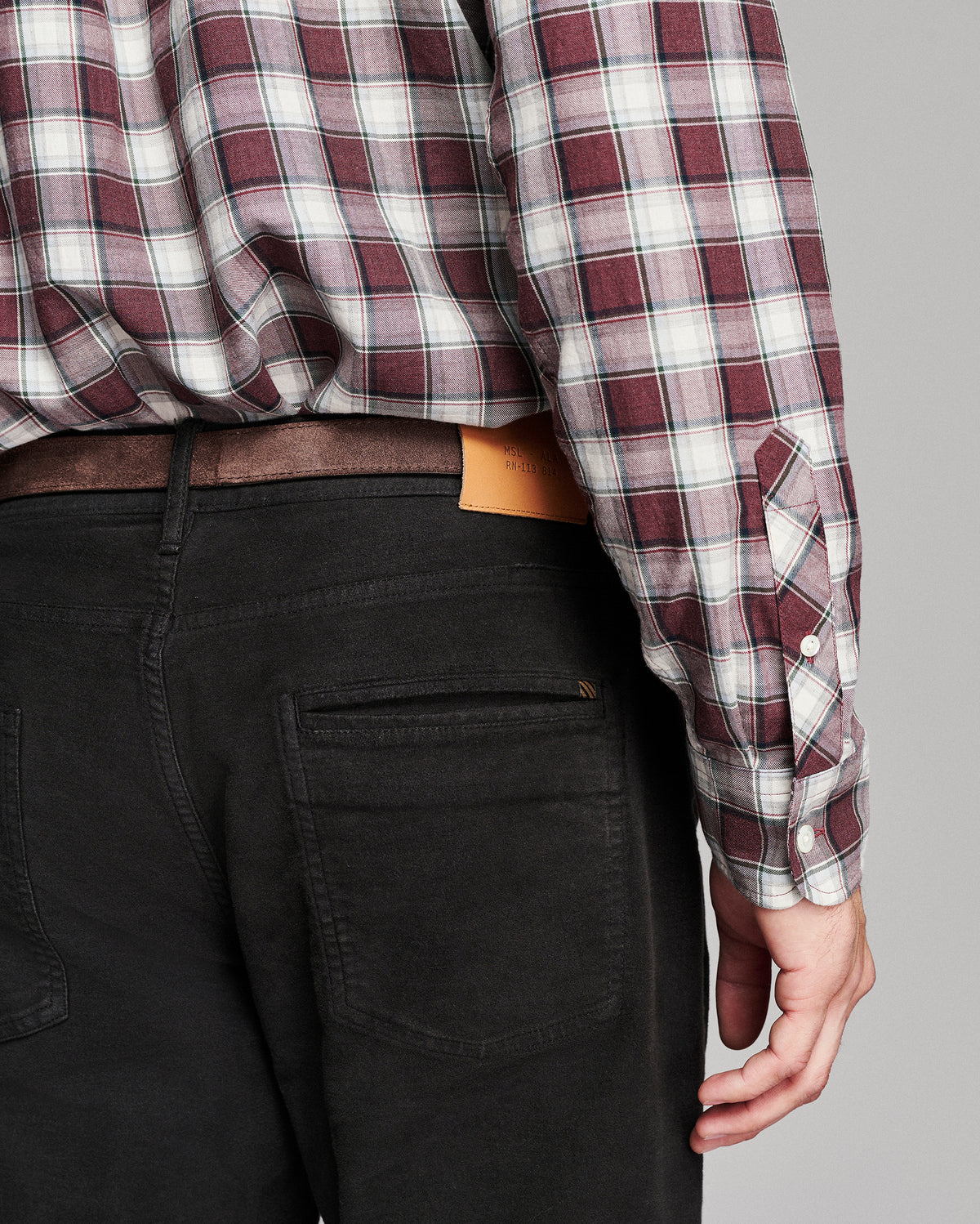 Close-up back view of a person wearing a burgundy, white, and gray plaid long-sleeve button-up shirt tucked into black jeans with a brown belt.