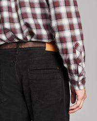 Close-up back view of a person wearing a burgundy, white, and gray plaid long-sleeve button-up shirt tucked into black jeans with a brown belt.