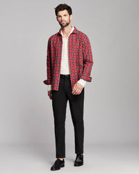 A man modeling a red and black plaid button-down shirt over a white collared shirt, paired with black trousers and black leather shoes, against a solid gray studio background.