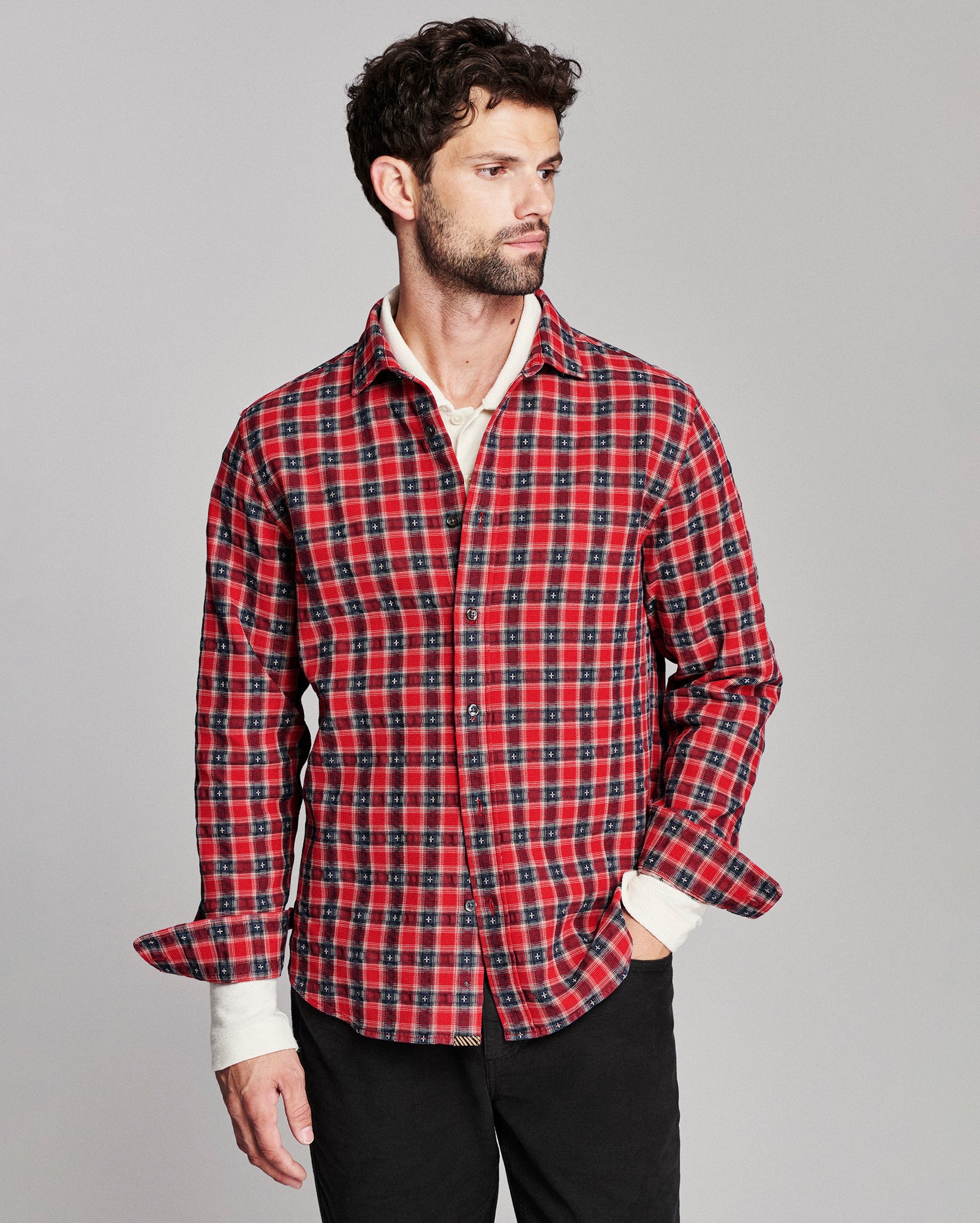 A man wearing a red and black plaid button-down shirt over a white undershirt, paired with black pants, photographed against a solid gray studio background.