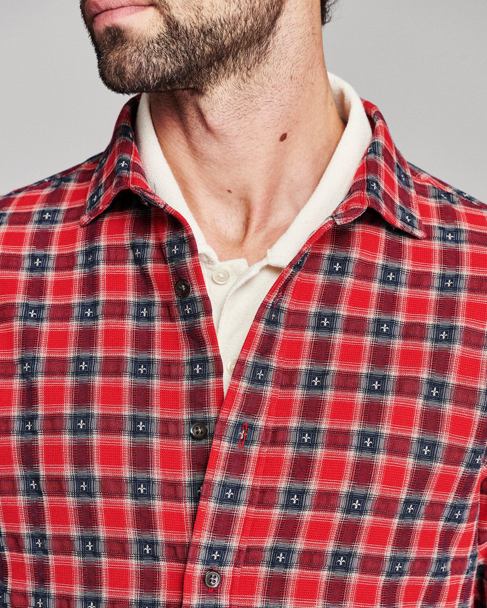Close-up of a man wearing a red and navy plaid button-up shirt over a cream polo shirt, cropped from the chin down against a light gray background.