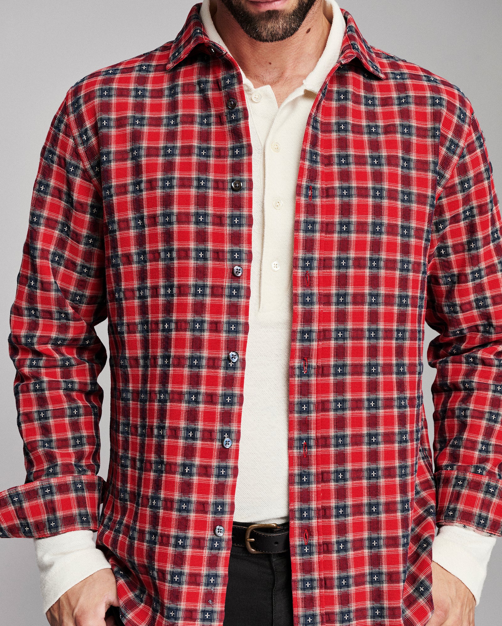 A man wearing a red and navy blue plaid button-down shirt over a cream collared shirt, shown from the chin down against a neutral gray background.