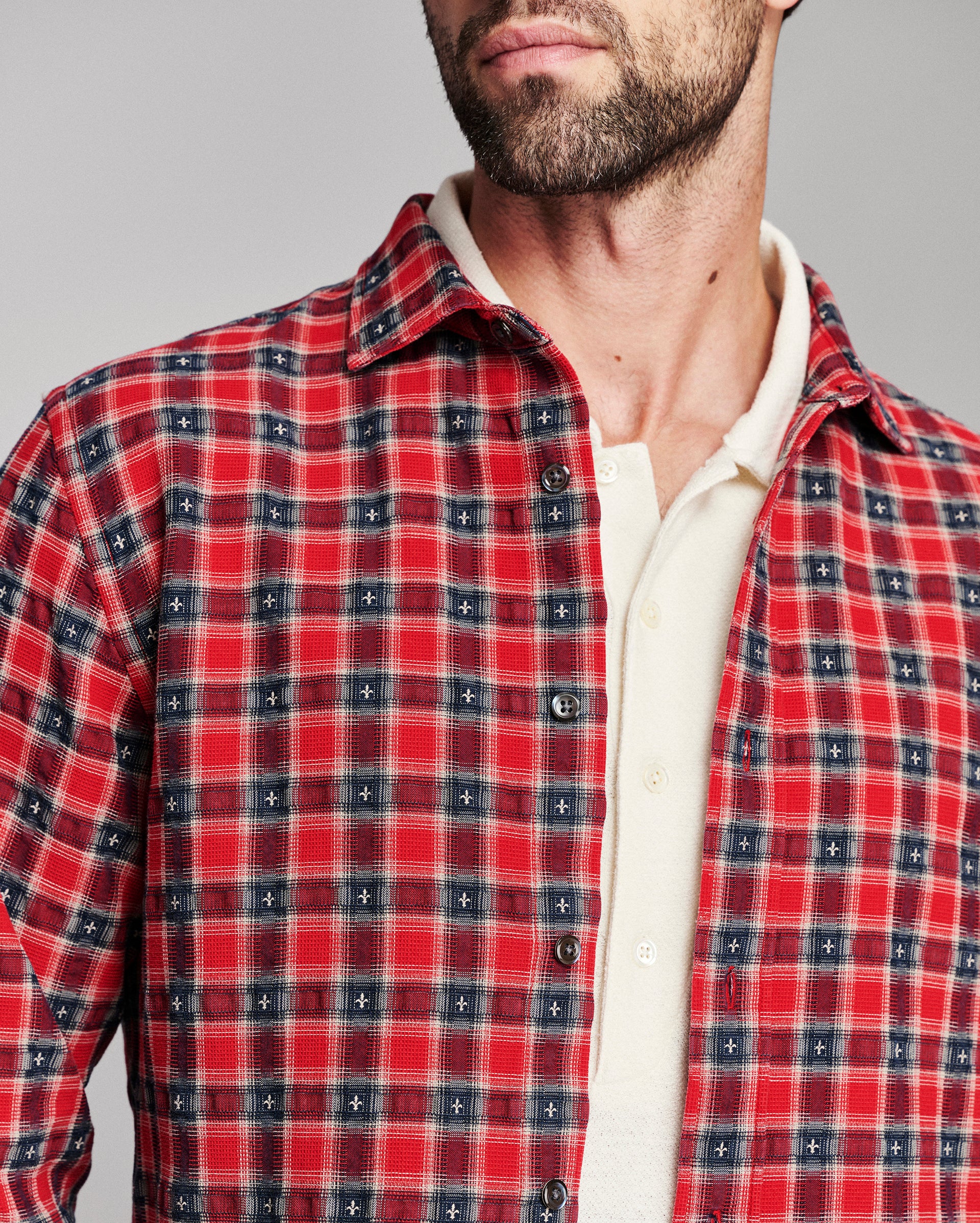 A close-up of a man wearing a red and navy plaid button-down shirt over a cream collared undershirt, against a plain gray studio background.