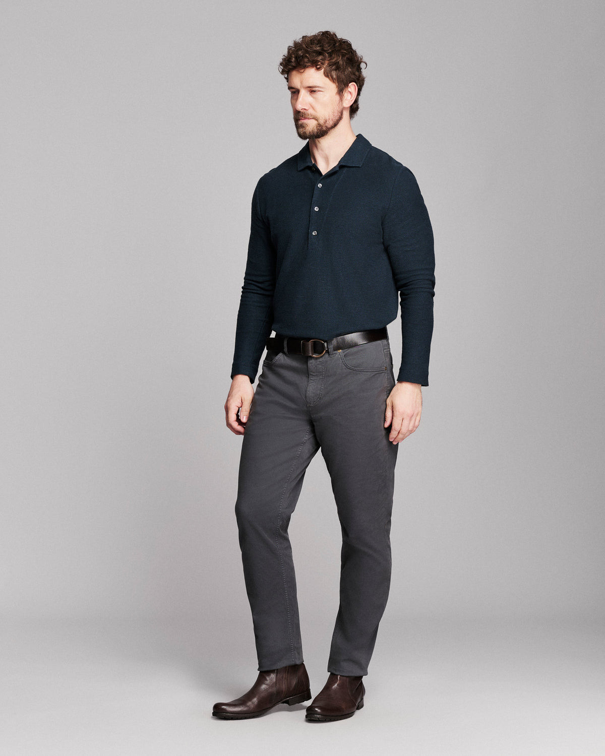 A man modeling a dark navy long-sleeve polo shirt, charcoal gray dress pants, a black belt, and dark brown leather dress shoes, standing against a neutral gray studio background.