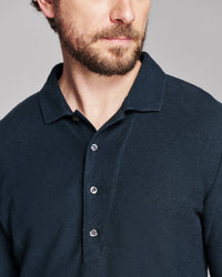 Close-up of a man wearing a dark navy blue textured polo shirt with a three-button placket, shown against a neutral gray background.
