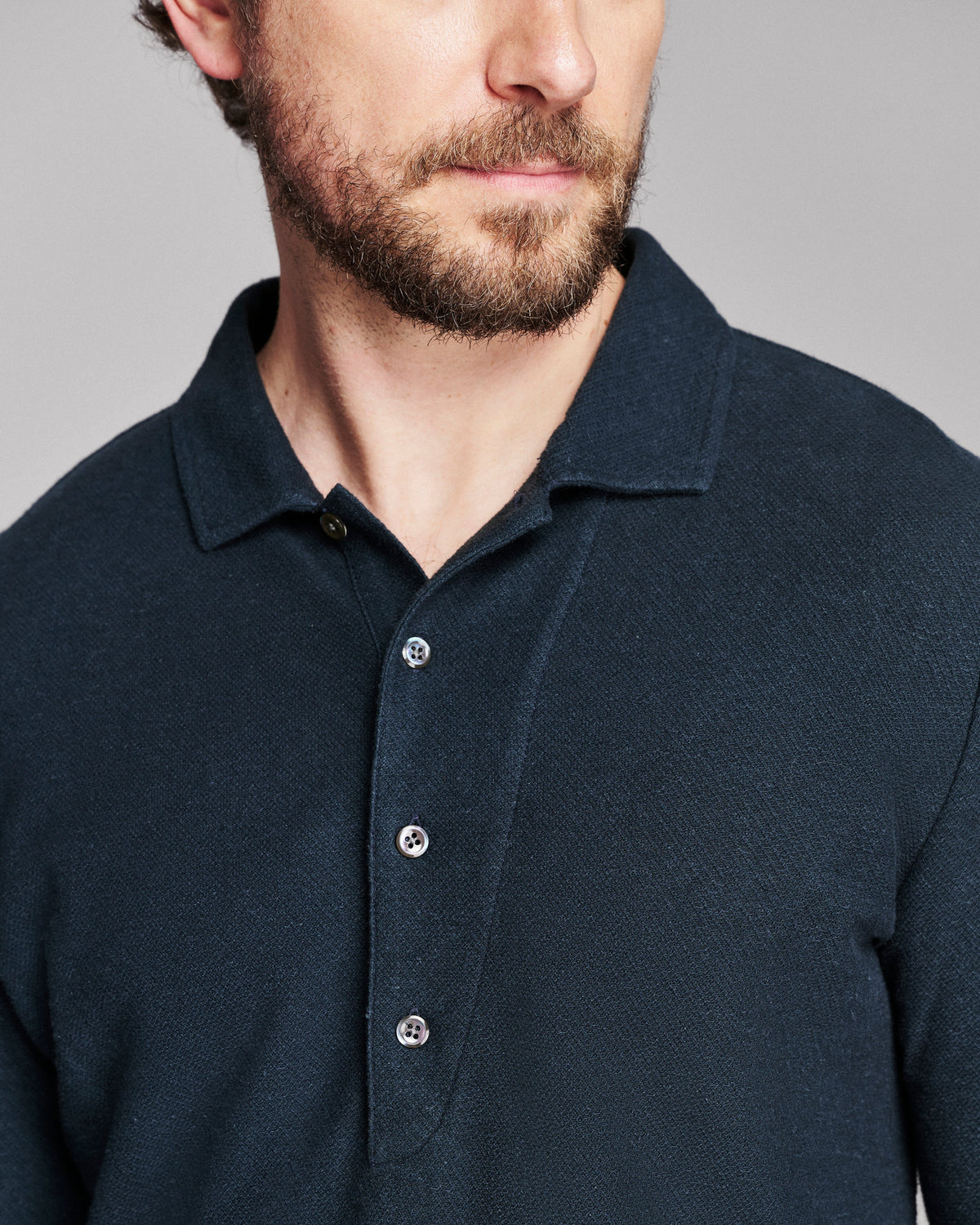 Close-up of a man wearing a dark navy blue textured polo shirt with a three-button placket, shown against a neutral gray background.