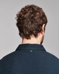 Back view of a man with curly brown hair wearing a dark navy blue collared shirt against a solid gray background.