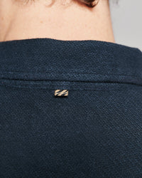 Close-up of a person's neck and upper back wearing a dark navy textured knit top, featuring a small gold-tone striped metal brand pin on the collar.