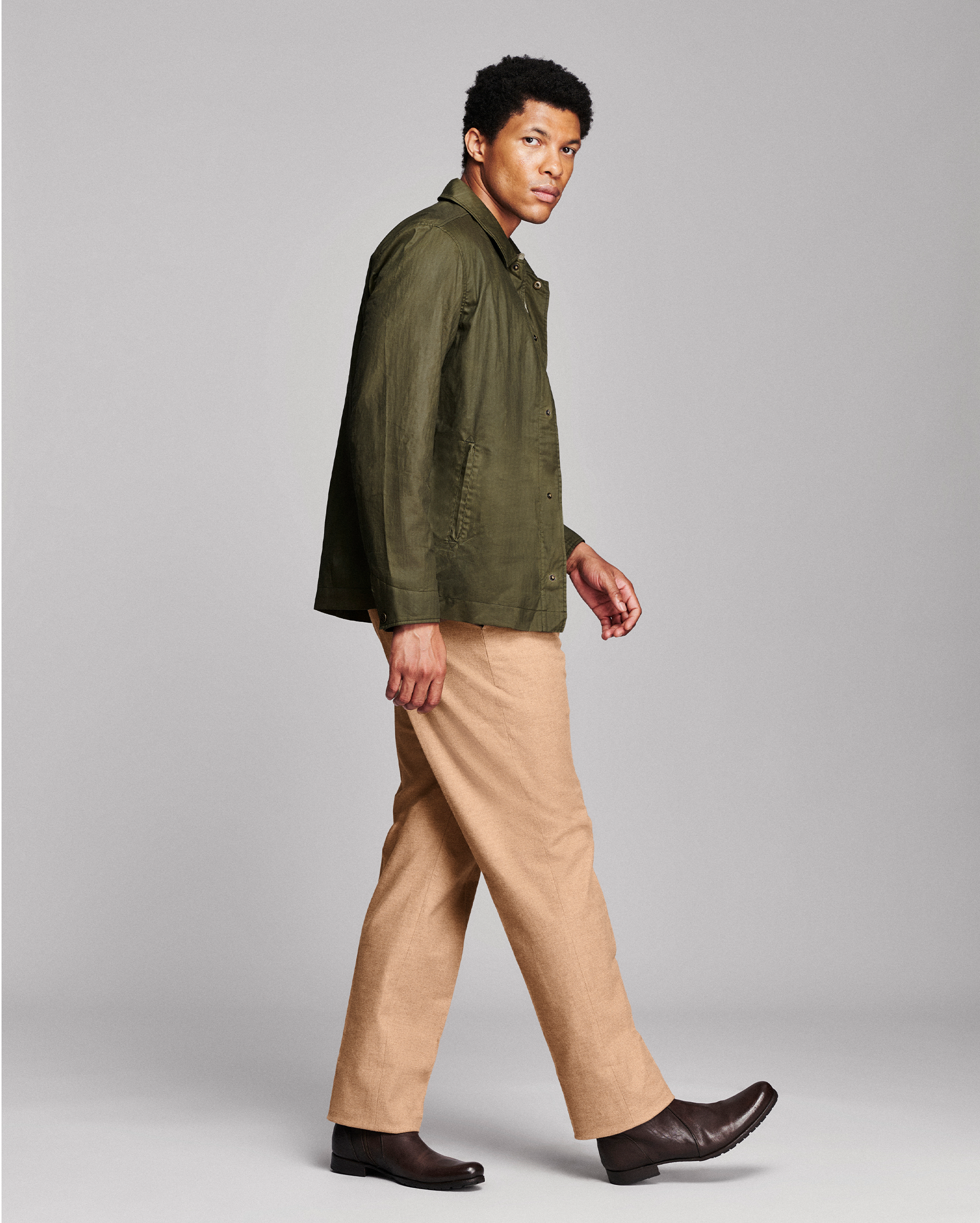 A man wearing an olive green button-up shirt, tan trousers, and dark brown leather shoes, captured in a side-profile walking pose against a solid light gray studio background.