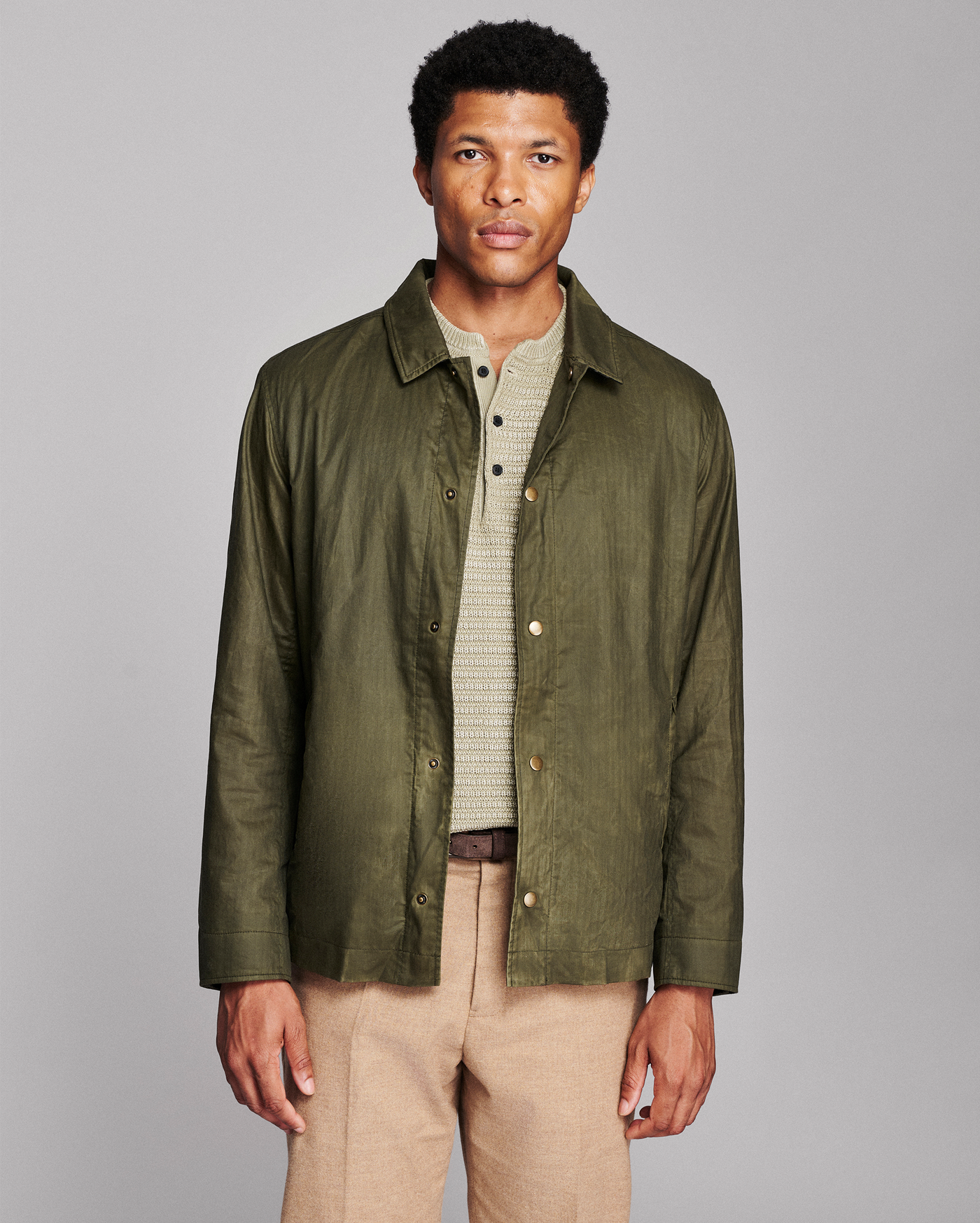 A man wearing an olive green lightweight button-up shirt jacket over a beige knit henley shirt and tan trousers, standing against a solid gray studio background.