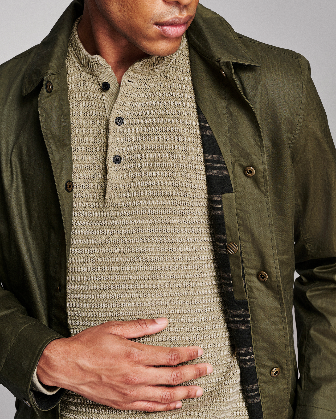 A close-up of a person wearing an olive green jacket over a beige ribbed knit henley shirt, with a dark striped inner layer partially visible.