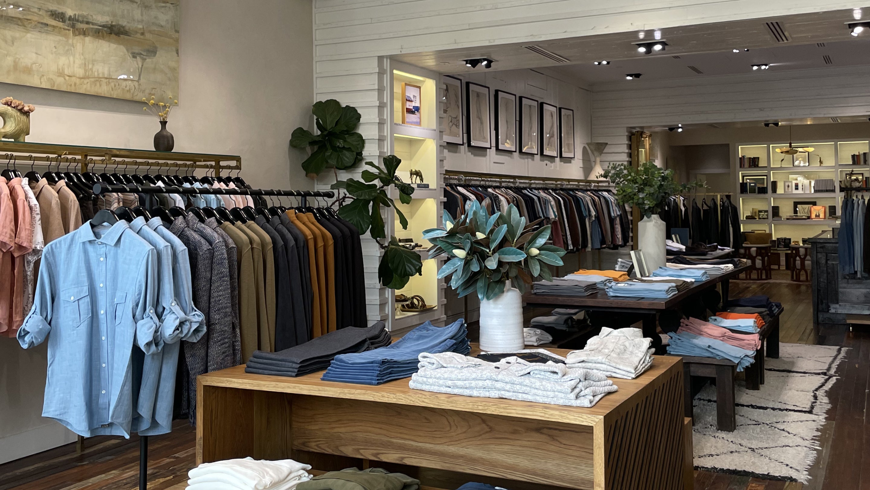 A well-lit clothing boutique interior showing racks of shirts, folded garments on tables, and decorative plants. A light blue button-up shirt is prominently displayed on a clothing rack on the left.