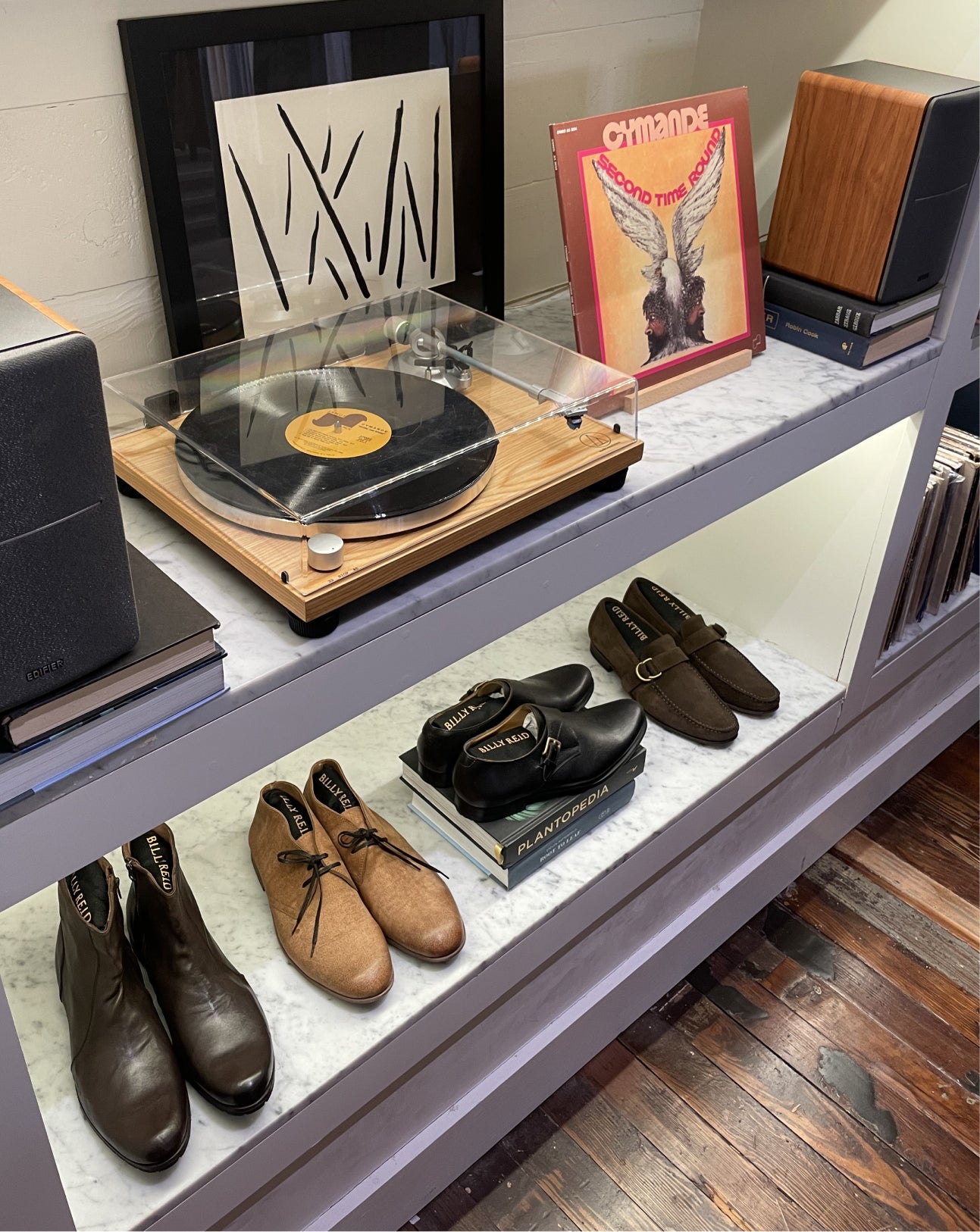 A styled interior shelf display featuring a wooden turntable with a vinyl record under a clear cover, framed wall art, a record sleeve, stacked books, and multiple pairs of shoes including brown leather boots, tan suede shoes, and black loafers.
